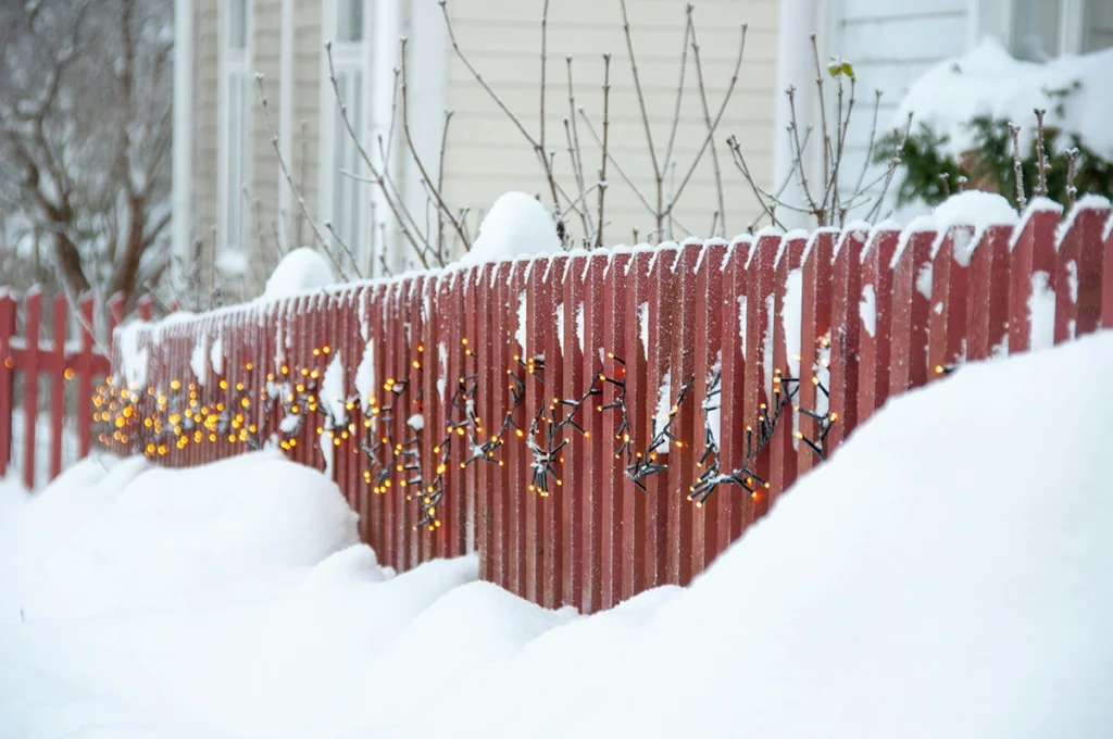 Red wooden fence in Ontario winter covered with snow and holiday lights, showing seasonal fence maintenance tips.